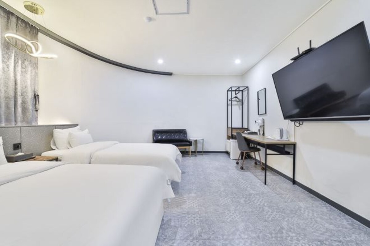 A modern hotel room with two beds, a desk, a chair, a flat-screen TV, and a small seating area.