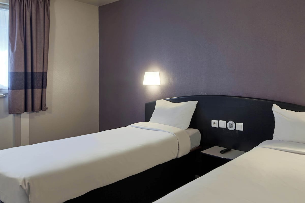 Habitación de hotel con dos camas individuales, una mesita de noche, una lámpara y una ventana con cortinas.