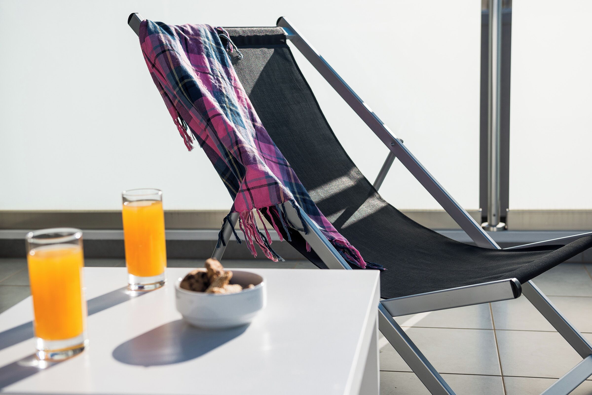 A sunlit balcony with a black lounge chair, a plaid blanket, a white table, and two glasses of orange juice.