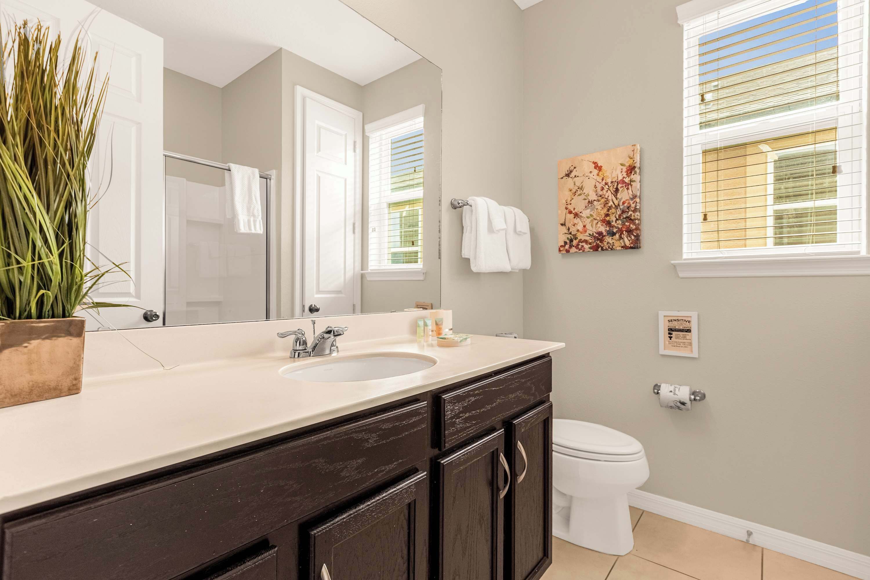 A bathroom with a large mirror, a sink, dark cabinets, a toilet, and a window with blinds.