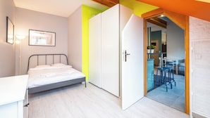 Une chambre à coucher comprenant un lit, une armoire et donnant sur un coin repas avec des tabourets.