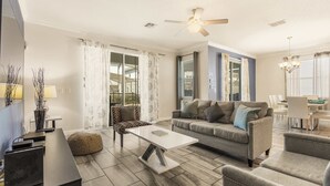 A modern living room with a sofa, coffee table, and ottoman, a dining area with a table and chairs, and a ceiling fan.