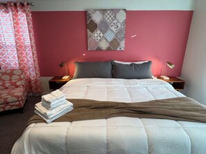 A neatly made bed with a white comforter and pillows, a bedside lamp, a nightstand with a lamp, and a patterned wall hanging.