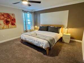 A bedroom with a bed, two bedside lamps, a ceiling fan, a window with curtains, and a painting on the wall. - Modern 6BR Villa Kissimmee | Perfect for 14 Guests (Kissimmee)