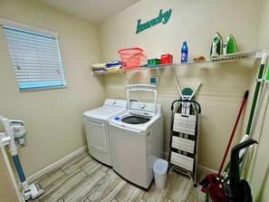 Une buanderie équipée d’un lave-linge, d’un sèche-linge, d’une étagère contenant des produits de lessive et d’un balai.