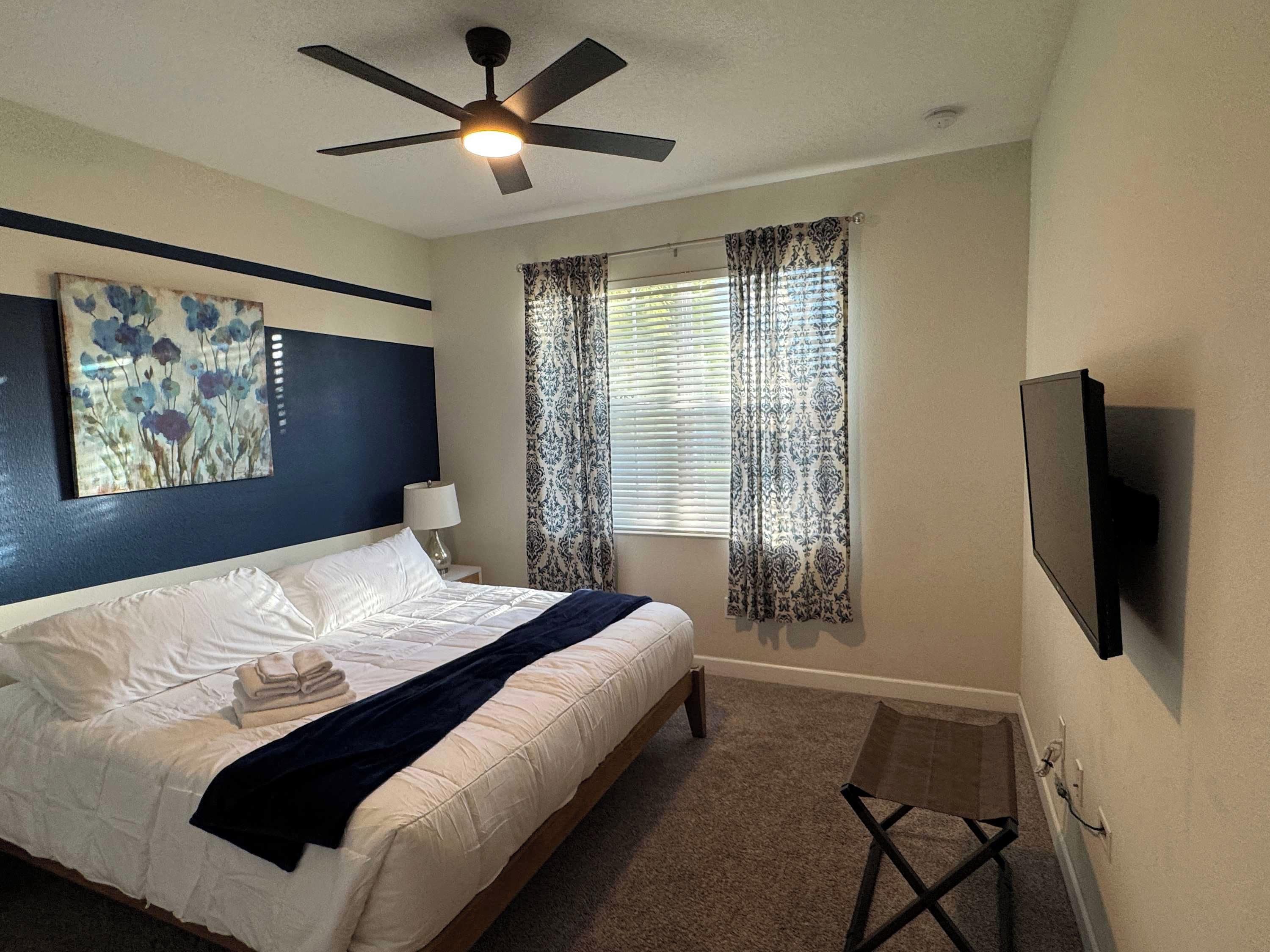 Habitación de hotel con cama, ventilador de techo, ventana con cortinas estampadas y televisor de pantalla plana montado en la pared.