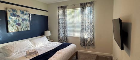 Habitación de hotel con cama, ventilador de techo, ventana con cortinas estampadas y televisor de pantalla plana montado en la pared.