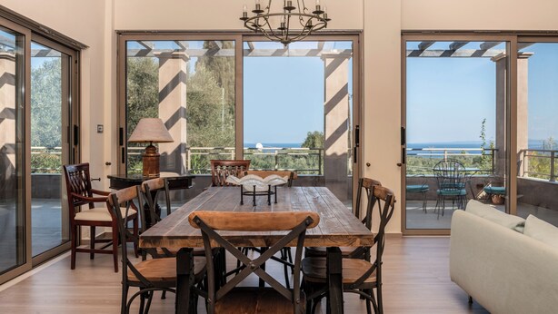 Àmplia zona de dinar amb taula i cadires de fusta, un penjallum i grans portes corredisses de vidre que donen a un pati exterior amb vista al mar.