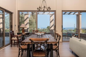 A spacious dining area with a wooden table and chairs, a chandelier, and large sliding glass doors leading to an outdoor patio with a view of the sea. - Astarte Villas - Stilvi 4 Bedroom Private Villa With Pool (RV3V+9Q3 Zakynthos, Greece)