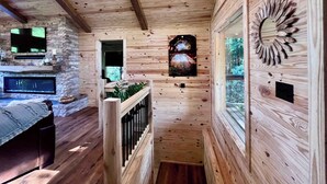 Interior de una cabaña de troncos con chimenea de piedra, televisor montado y techo de madera.