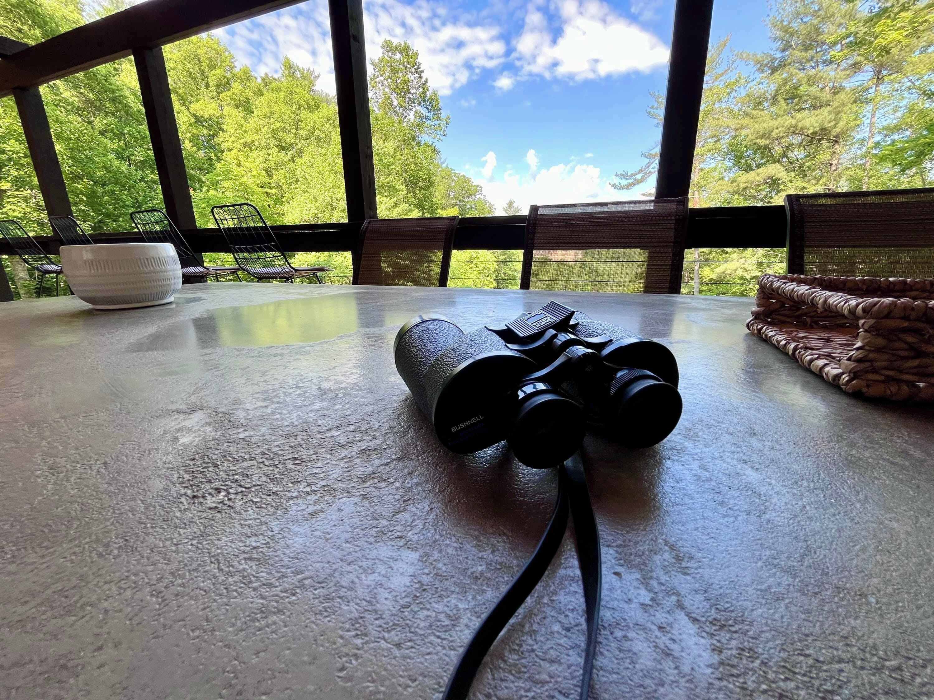 A telescope on a table with a view of trees and sky through large windows.