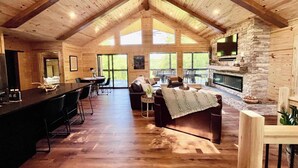 Un salón amplio con techo alto, vigas de madera, chimenea de piedra y ventanas grandes que ofrecen vistas al exterior.