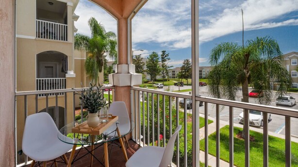 A balcony with a glass table, two white chairs, a potted plant, and a view of a residential street.
