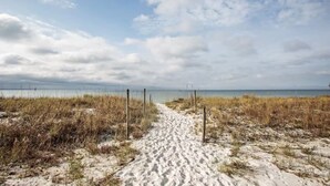 Ein sandiger Küstenpfad führt zum Meer, begleitet von einem Zaun auf einer Seite und Gras auf der anderen.