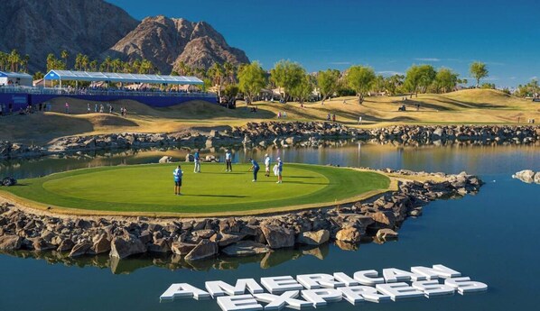 Ein Golfplatz mit einem Wasserspiegel, einem Berg im Hintergrund und einem großen Schild mit der Aufschrift 'AMERICAN EXPRESS'.