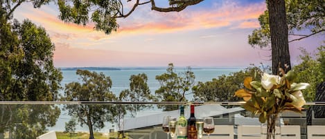Ein Tisch im Freien mit Blick auf Meer und Bäume, eine Weinflasche und ein Blumenstrauß.