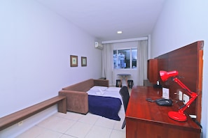 A hotel room with a bed, a desk with a red lamp, a telephone, and a window with curtains.