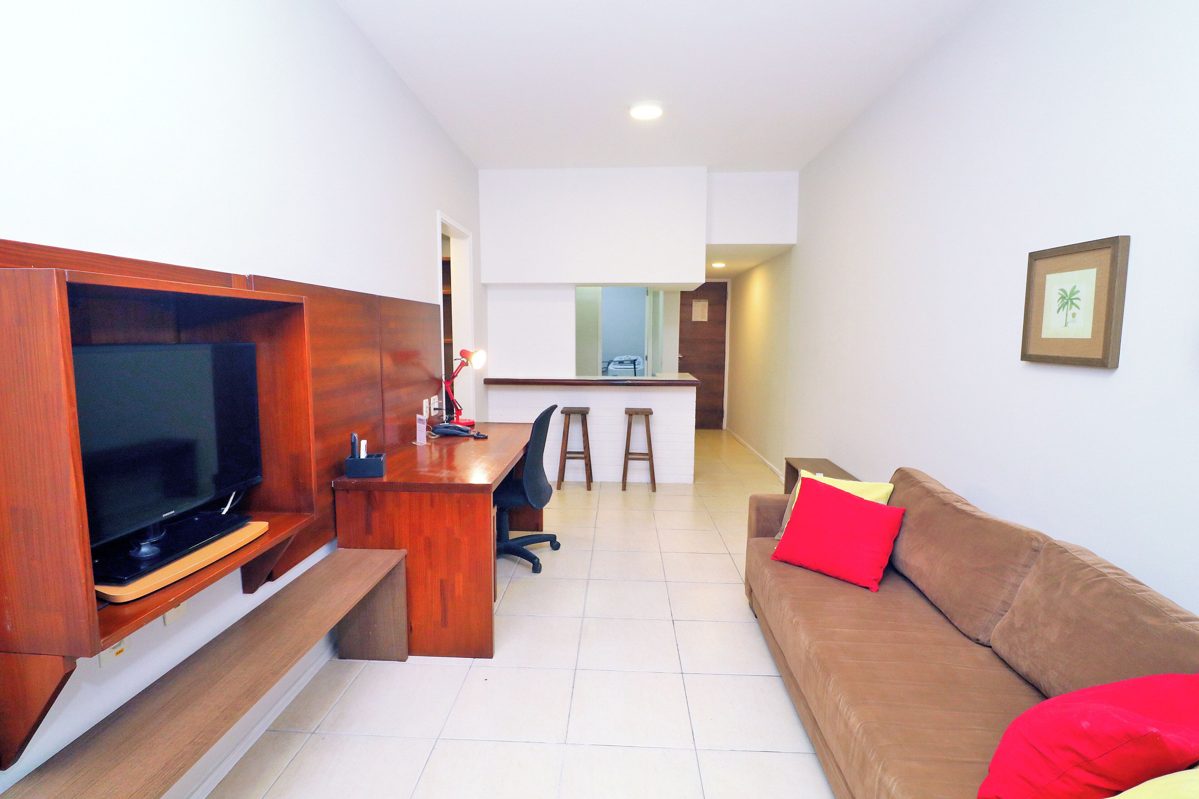 Una sala de estar moderna con un mueble de entretenimiento de madera, un sofá marrón con cojines rojos y una cocina al fondo.