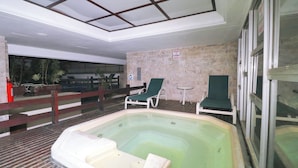 Ett inomhusområde med en simbassäng, ett bubbelbad, två gröna solstolar, ett trädäck och en stenmur.