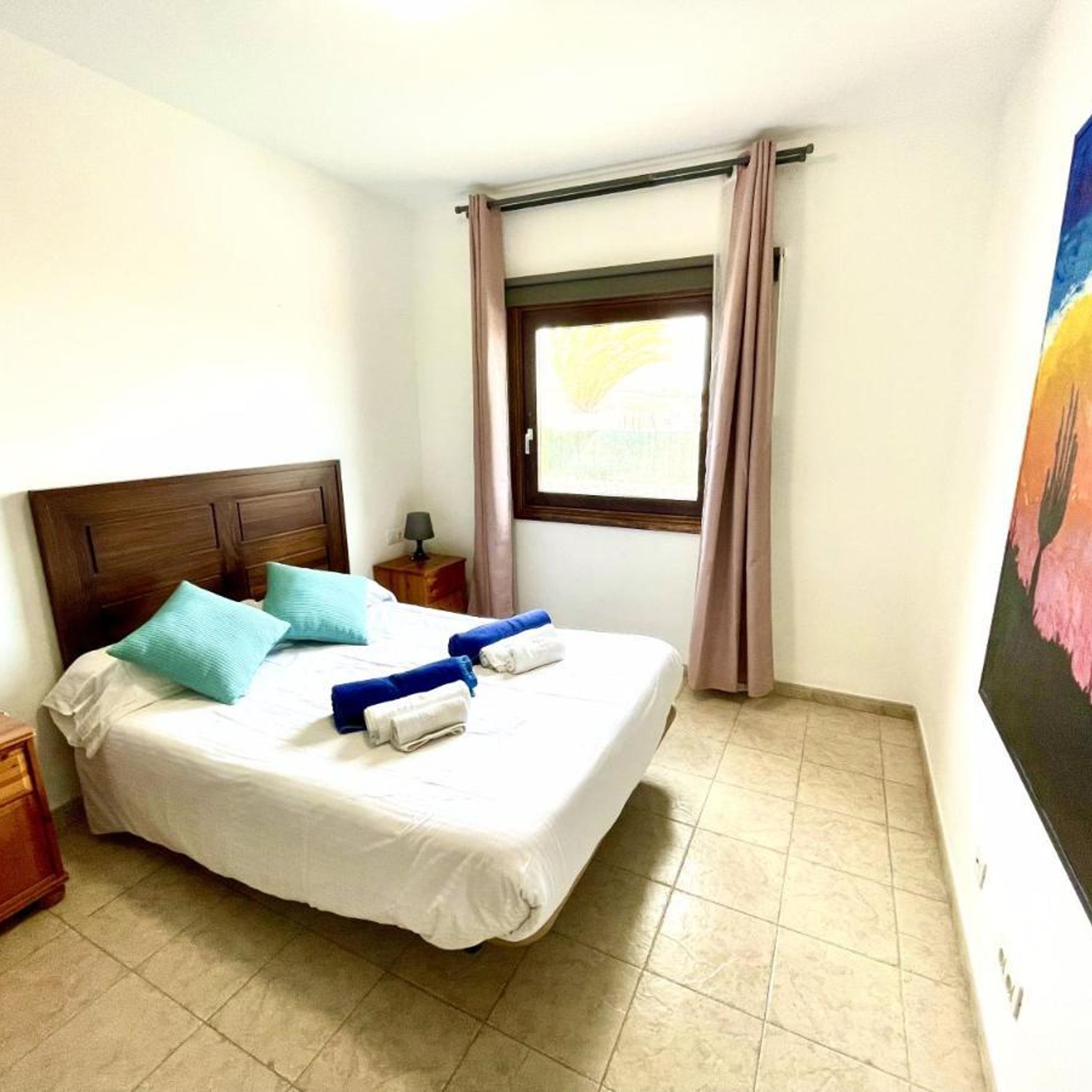 A neatly made bed with pillows, a nightstand, a lamp, a window with curtains, and a painting on the wall.