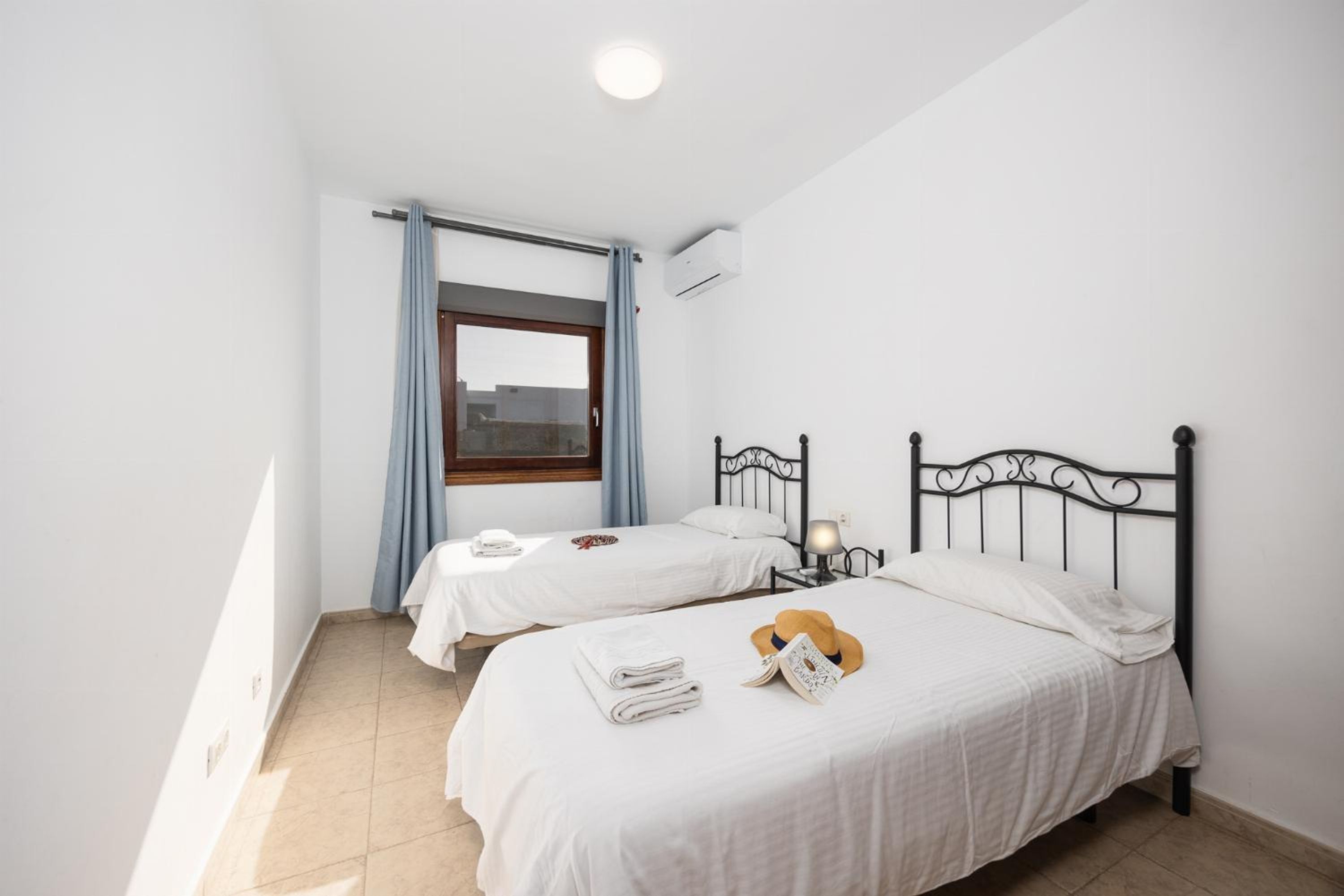 A bedroom with two beds, a window with curtains, an air conditioner, and a bedside table with a lamp.