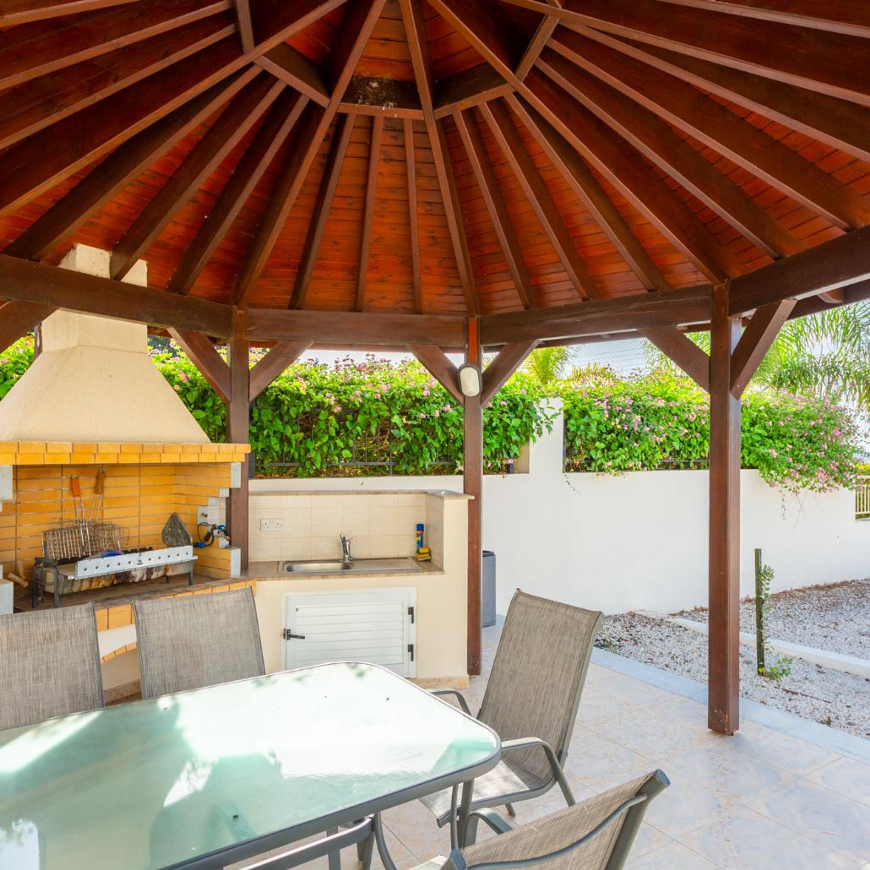 An outdoor patio with a wooden gazebo, a glass table, and a grill.