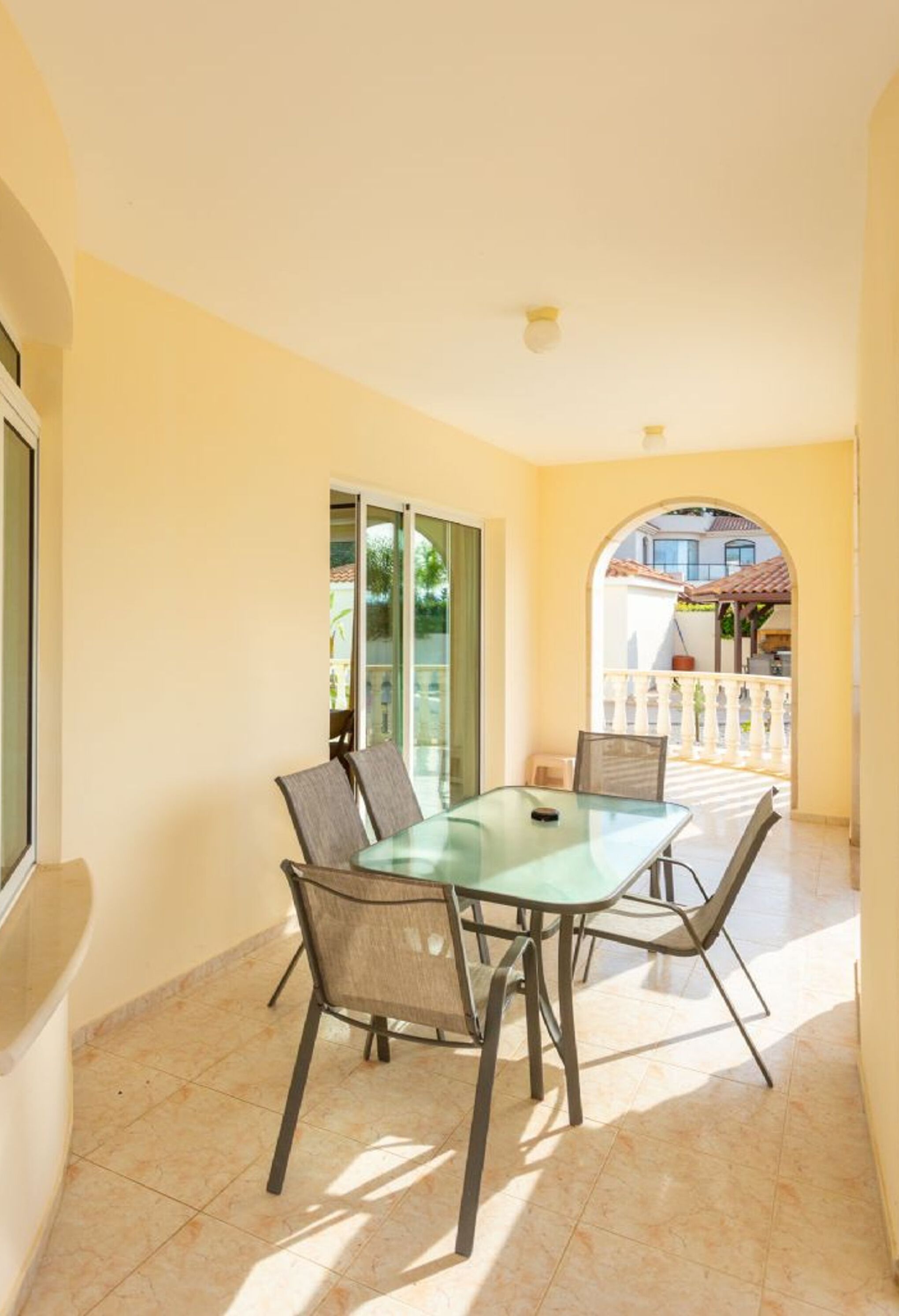 A sunlit patio with a glass table and chairs, a sliding glass door leading to a balcony, and a view of a residential area.