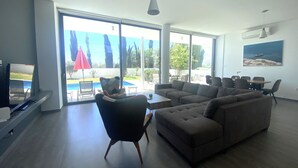 A spacious living room with a large sectional sofa, a coffee table, and a dining area with chairs and a table. There is a sliding glass door leading to an outdoor area with a pool and umbrellas.