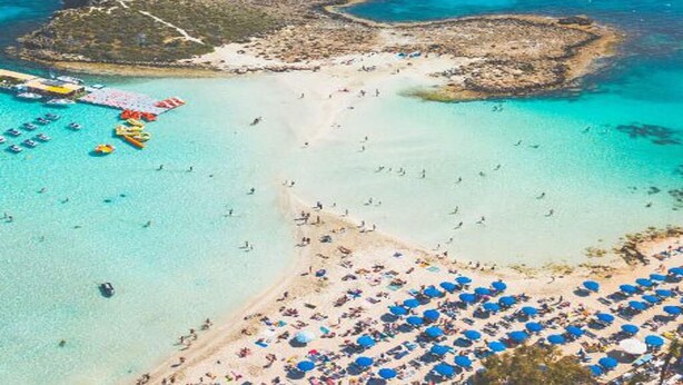 An aerial view of a beach with clear turquoise waters, a sandy shore, and numerous umbrellas.