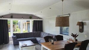 En rummelig opholdsstue med en stor sofa, et spisebord og et sofabord. Der er et vindue med persienner og en pendellampe.