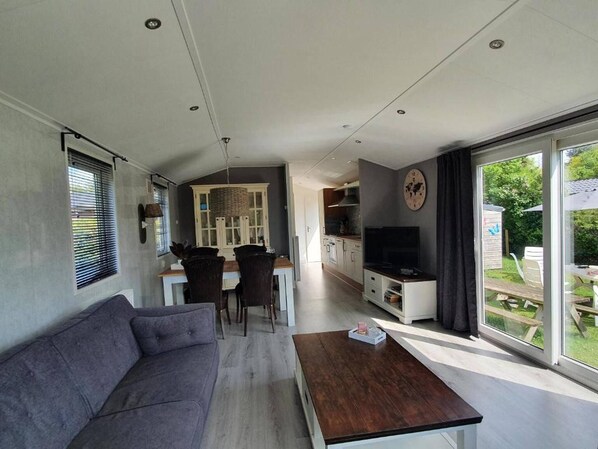 A modern living room with a sofa, a coffee table, and a TV. There is a dining area with a table and chairs, and a kitchen in the background.