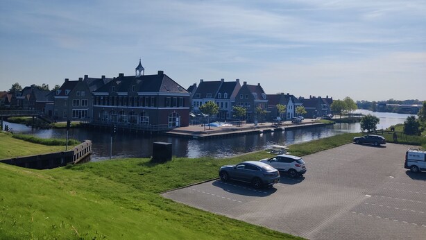 A canal-side building with a waterway, parked cars, and a grassy area. - Paloma Chalet with Sauna - Relaxing Nature Escape by Lauwersmeer Park (Anjum)