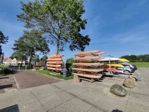 A row of canoes and kayaks parked on a paved area with a tree and buildings in the background. - Paloma Chalet with Sauna - Relaxing Nature Escape by Lauwersmeer Park (Anjum)