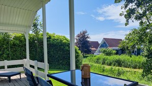 A covered outdoor seating area with a glass table and chairs, surrounded by greenery and a view of houses. - Paloma Chalet with Sauna - Relaxing Nature Escape by Lauwersmeer Park (Anjum)