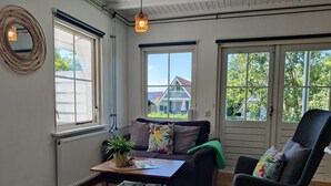A cozy living room with a sofa, a chair, a coffee table, and a window overlooking a house. - Paloma Chalet with Sauna - Relaxing Nature Escape by Lauwersmeer Park (Anjum)