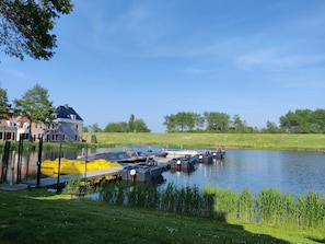 A calm lake with boats docked, a grassy bank, and a house in the background. - Paloma Chalet with Sauna - Relaxing Nature Escape by Lauwersmeer Park (Anjum)