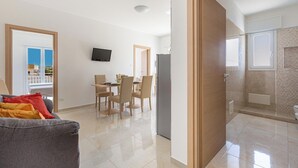 A modern living room with a grey sofa, a dining area with chairs, a TV mounted on the wall, and a bathroom with a shower and toilet. - 3952 Trilo di Annahouse by Barbarhouse (Porto Cesareo)