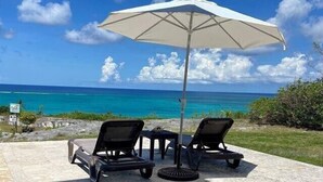 Een terras met twee loungestoelen en een grote parasol met uitzicht op een strand met turquoise water en een heldere lucht.