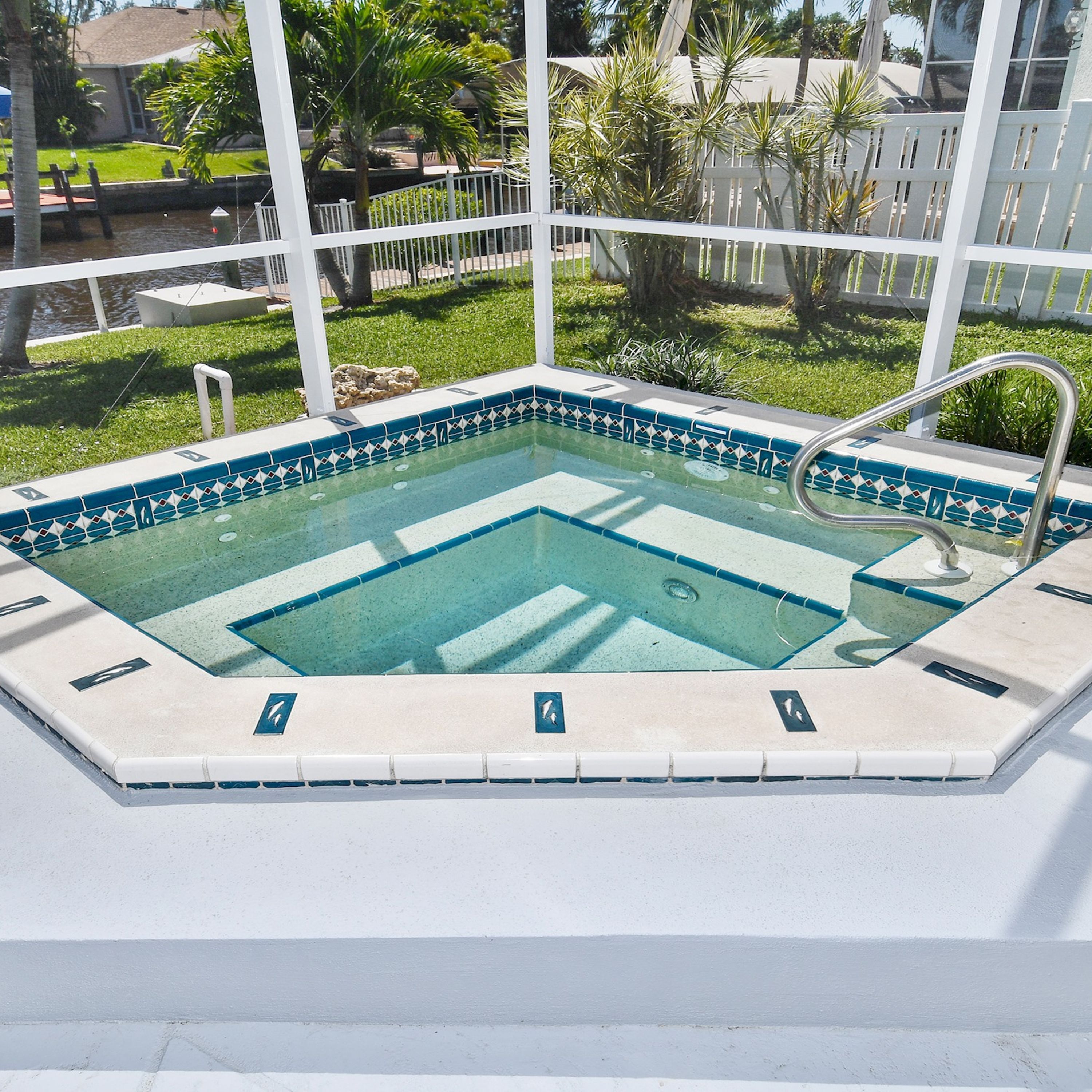 A rectangular hot tub with a tiled edge and a metal ladder, surrounded by a white fence and greenery.