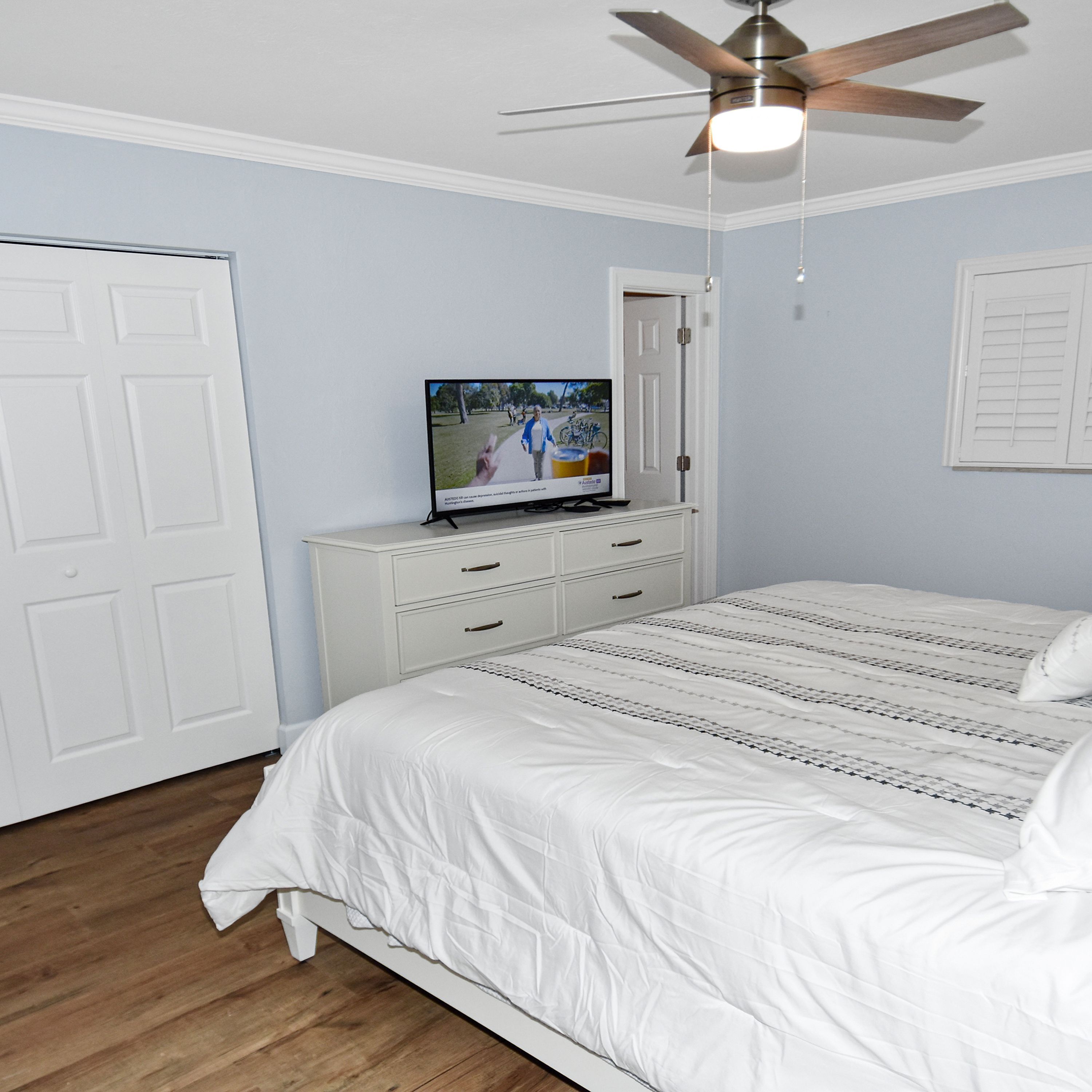 A bedroom with a bed, a TV on a dresser, a ceiling fan, and a door.
