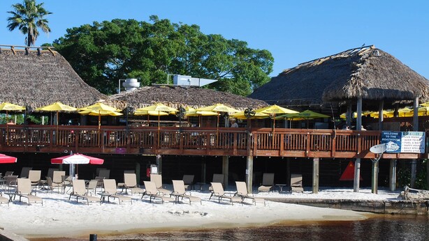 Un bar situé en bord de mer, doté d’un toit de chaume, de tables à l’extérieur et d’une terrasse en bois offrant une vue sur l’eau.
