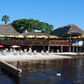 Un bar situé en bord de mer, doté d’un toit de chaume, de tables à l’extérieur et d’une terrasse en bois offrant une vue sur l’eau.