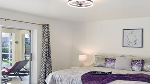 A bedroom with a bed, a nightstand, a lamp, a chair, and a view through a glass door. - Su Casa - XXL Pool Area Pool Spa 3 Bedroom 2 Bath (Cape Coral)