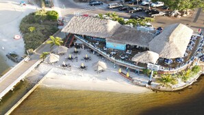 Vue aérienne d’un hôtel situé en bord de mer, avec une structure à toit de chaume, des espaces de détente en plein air et une jetée qui s’avance dans l’eau.