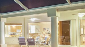 Un espace aménagé au bord de la piscine avec une table et des chaises, une cuisine visible à travers une porte ouverte, et une piscine dont le bord est revêtu de carreaux.