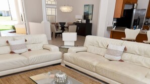 A modern living room with two white sofas, a glass coffee table, and a kitchen in the background.