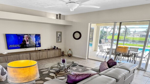 A modern living room with a grey sofa, a TV on the wall, and a view of the outdoors through large windows. - Timeout -south Facing With Large Terrace and Pool (Cape Coral)