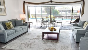 A living room with a grey sofa, a coffee table, and a view of a pool through large windows. - Belle Paradise - 3 3 Pool Boating Oasis of Calm (Cape Coral)