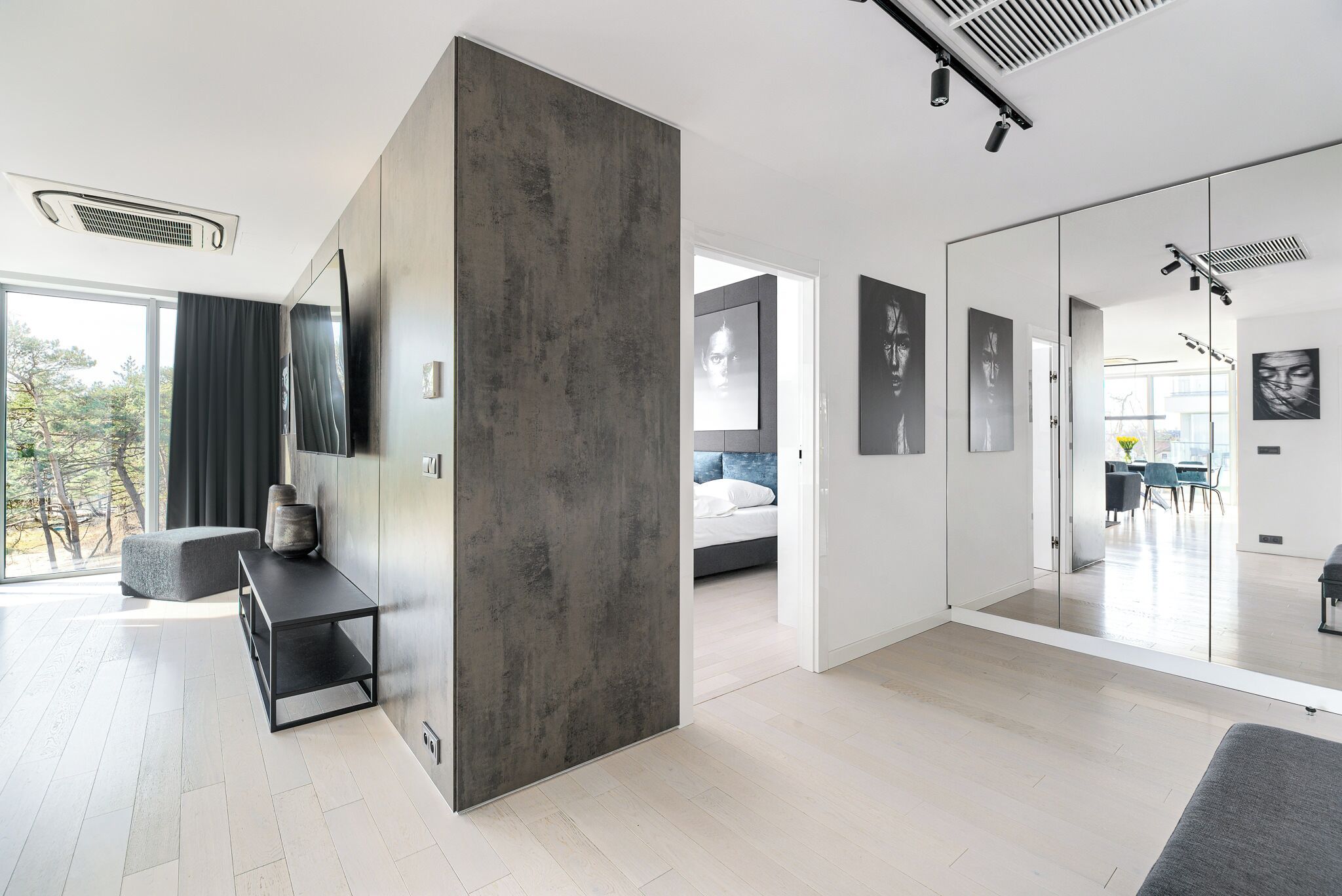 Una sala de estar moderna con un amplio separador gris, una mesa de centro negra y un sofá. En el fondo, hay una cama con ropa de cama blanca y un comedor con una mesa y sillas.