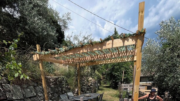 Una terraza de madera con pérgola, una zona de estar cubierta y un muro de piedra.